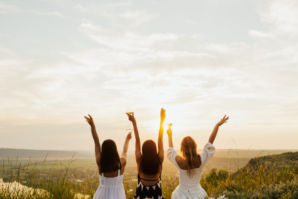 Three women raising glasses in a toast against a sunset backdrop, celebrating together outdoors.