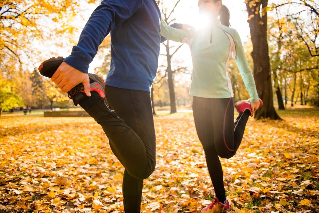Couple performing a stretching exercise outdoors in an autumn park with fallen leaves.
