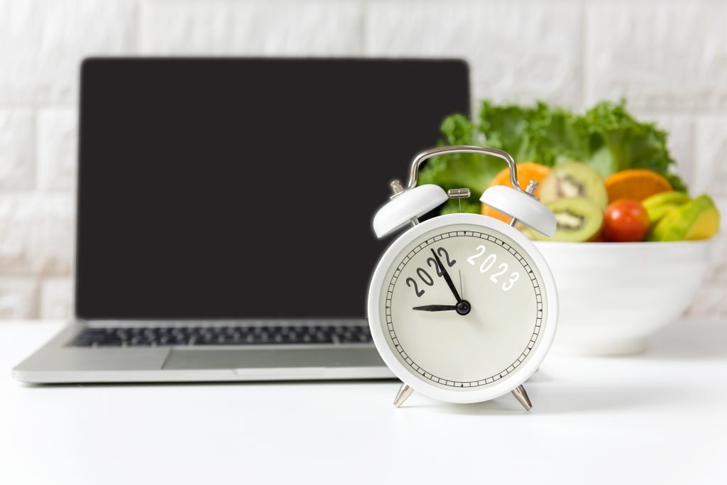 White alarm clock displaying the years 2022 and 2023 in front of a laptop and a bowl of fresh fruits and vegetables.