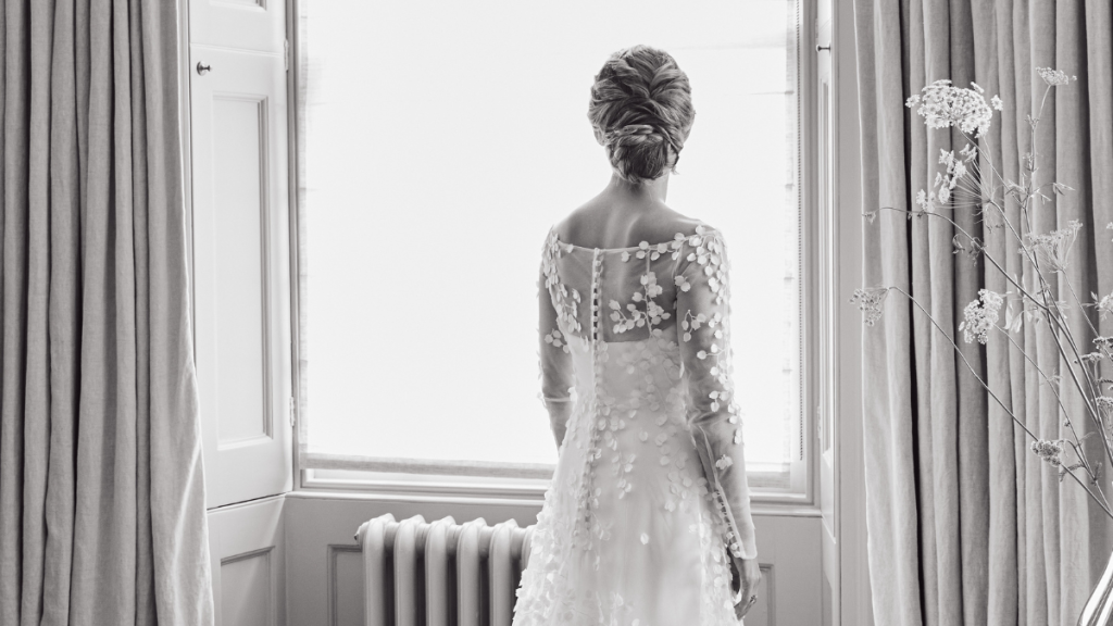 Bride wearing a white wedding dress with floral embellishments, standing by a window in a softly lit room.