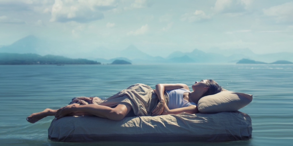 Individual lying on a floating bed in calm water, surrounded by mountains and clouds.