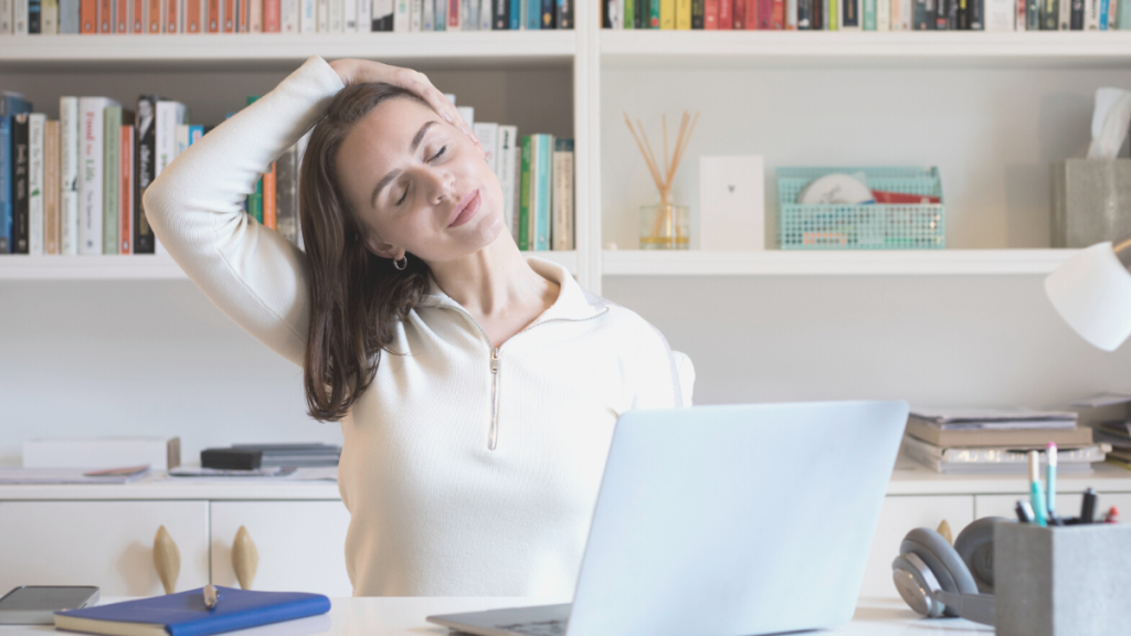 Woman performing a neck stretch while sitting at a desk with a laptop and books in the background.