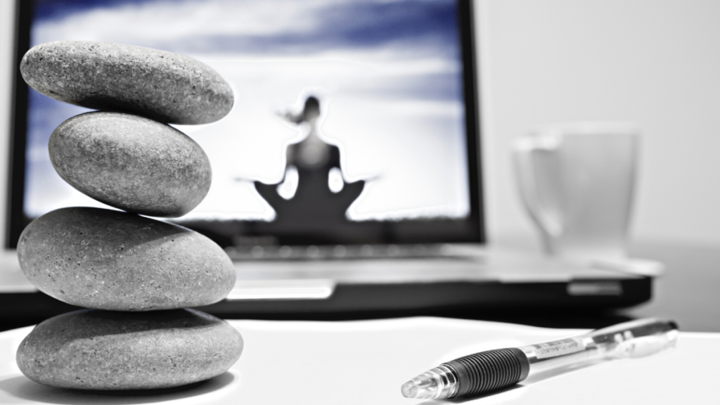 A balanced stack of four grey stones in front of a laptop displaying a meditative figure and a pen on a notepad.