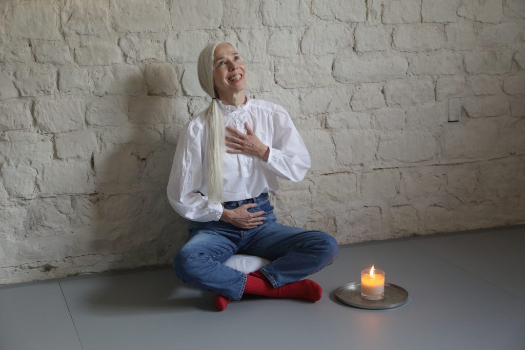 Woman with long silver hair wearing a white blouse, smiling and looking upwards against a textured stone wall.