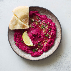 Beetroot dip served in a bowl with a slice of pita bread and a wedge of lime, garnished with seeds and herbs.