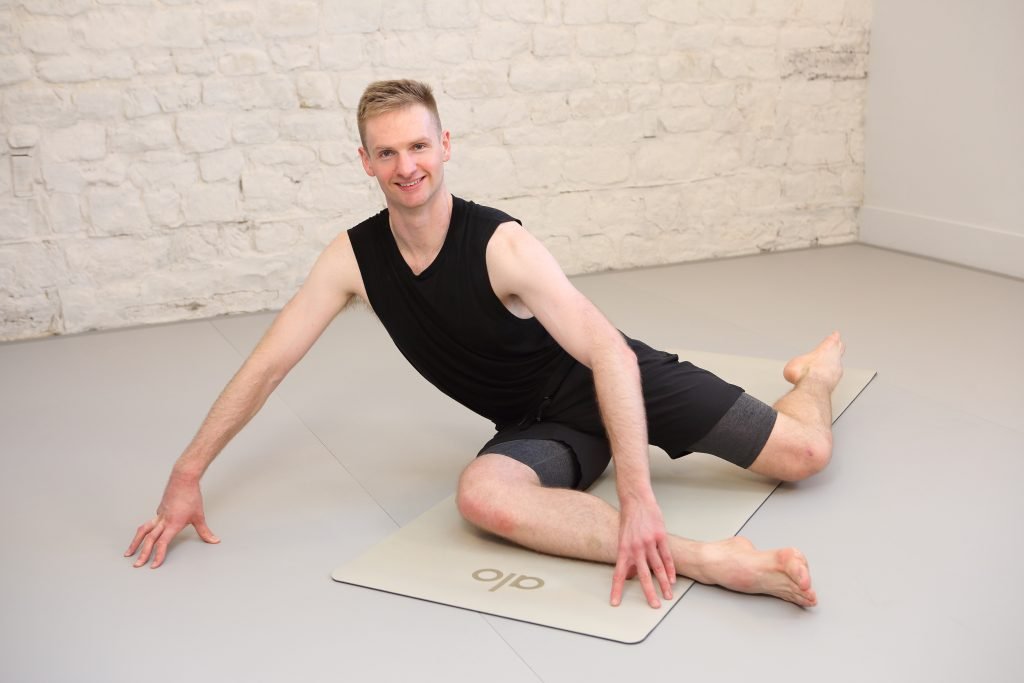 Man in a black sleeveless top and grey shorts performing a yoga pose on an Olo mat in a studio setting.