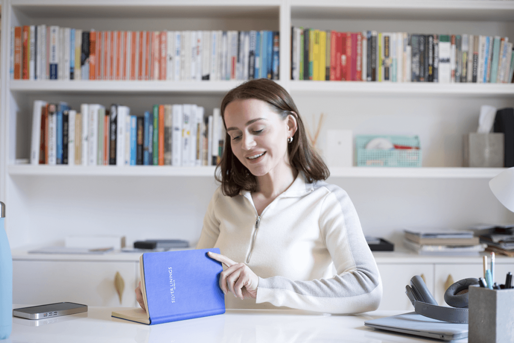 A woman in a light sweater interacting with a blue book titled 'Silverlining' while seated at a desk.