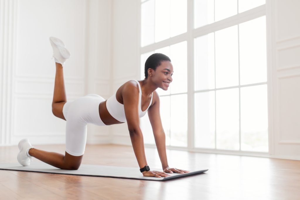Woman in white athletic wear performing an exercise on a yoga mat in a bright room with large windows.