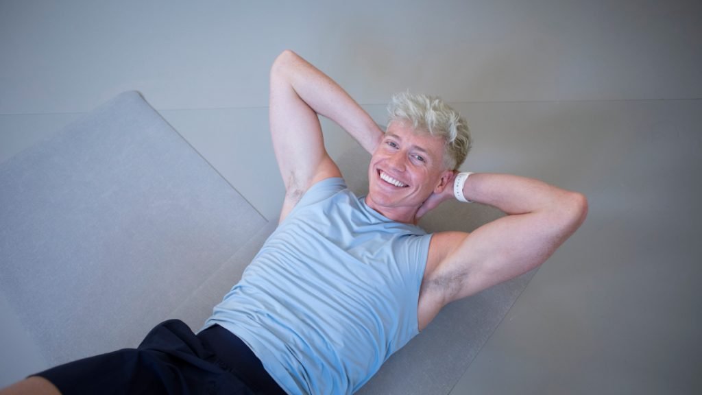 Man with blonde hair performing a sit-up on a mat, wearing a light blue sleeveless top and black shorts.