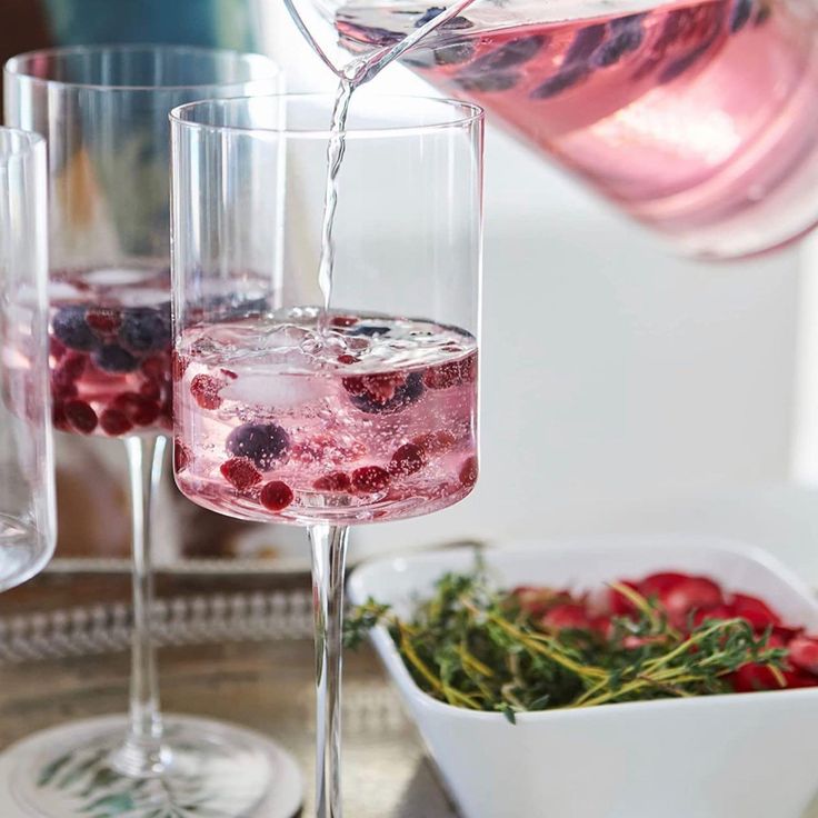 Pouring a pink beverage with berries into a clear glass, with additional berries and herbs in a bowl.