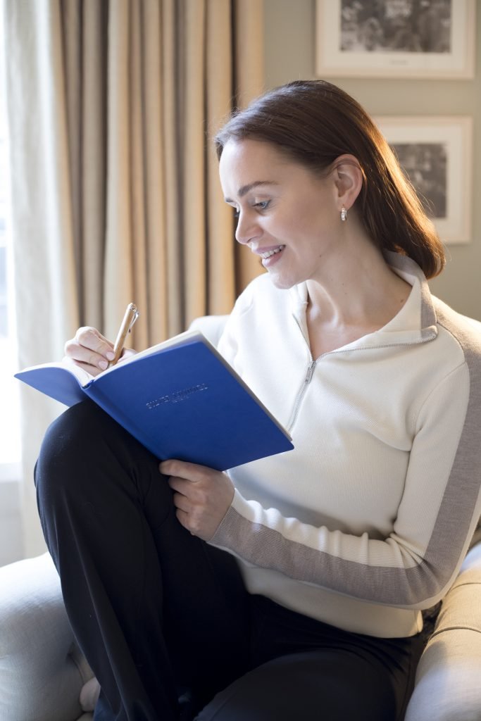 Woman sitting in a chair, writing in a blue notebook with a pen, wearing a white and grey top.