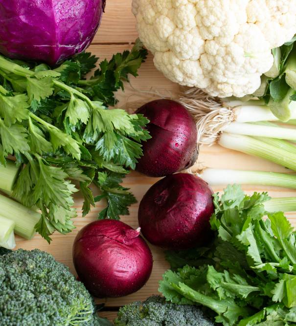 A selection of seasonal vegetables including broccoli, cauliflower, red onions, and celery arranged on a wooden surface.