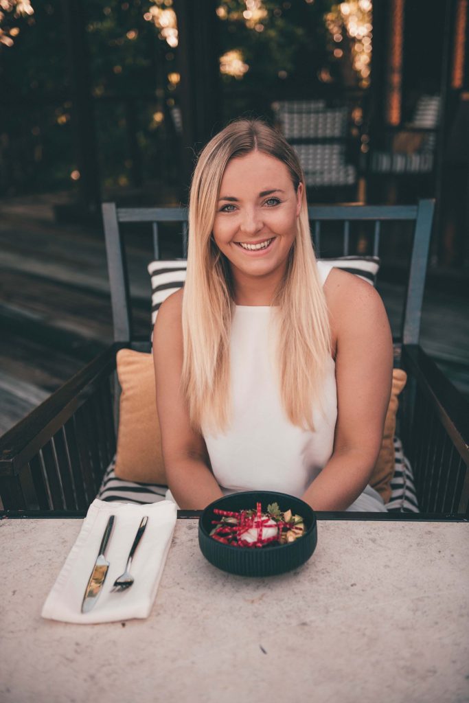 A woman with long blonde hair sitting at a table with a bowl of fruit and cutlery.