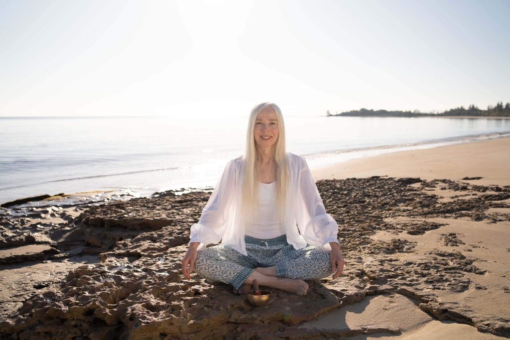 Catherine sitting cross-legged on the beach, smiling while meditating on a rocky shoreline.