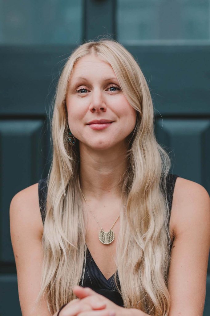 A woman with long blonde hair and a necklace, seated and looking directly at the camera.