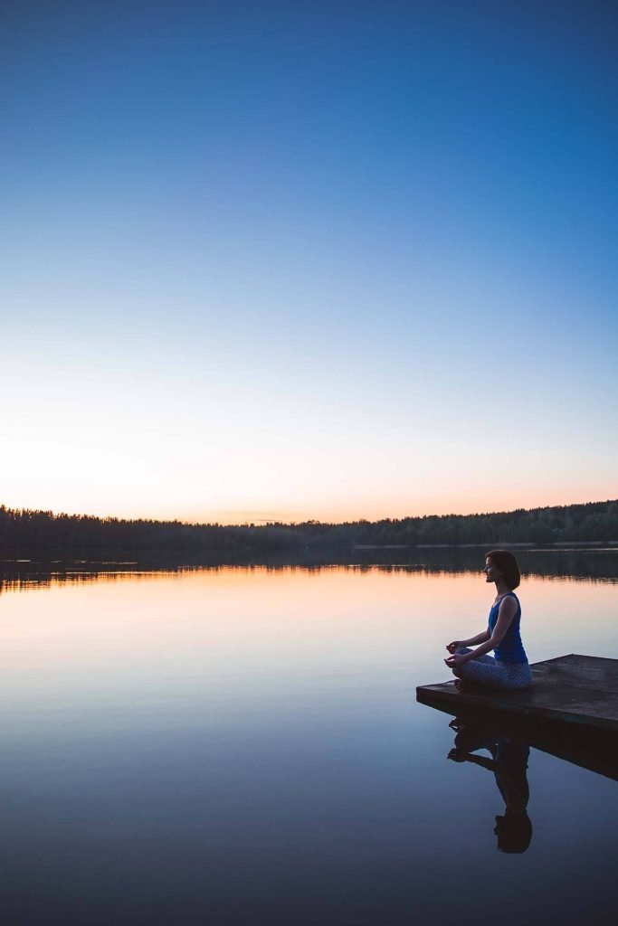Individual sitting cross-legged on a dock by a calm lake during sunset, reflecting the serene sky.