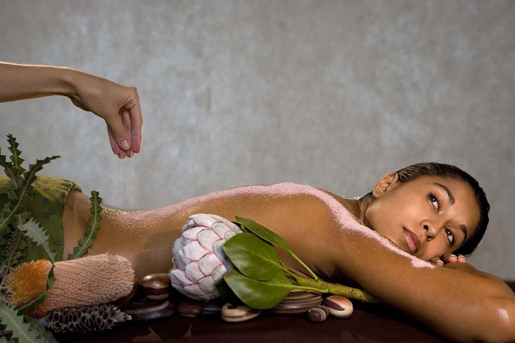Woman lying on a massage table with pink salt being sprinkled on her back, surrounded by stones and botanical elements.