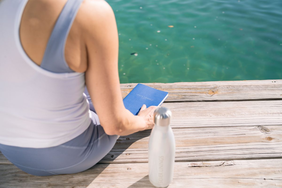 Individual seated on a wooden dock holding a blue notebook next to a silver water bottle by the water's edge.