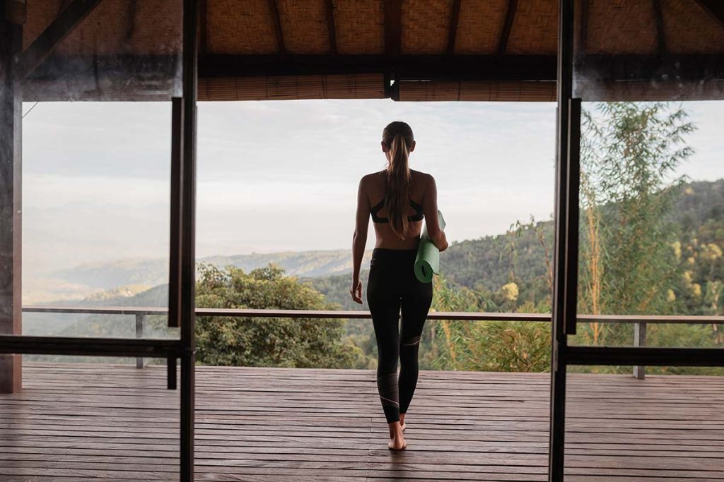 Young woman in sport clothes holding a rolled yoga mat, walking towards a balcony with a mountain view.