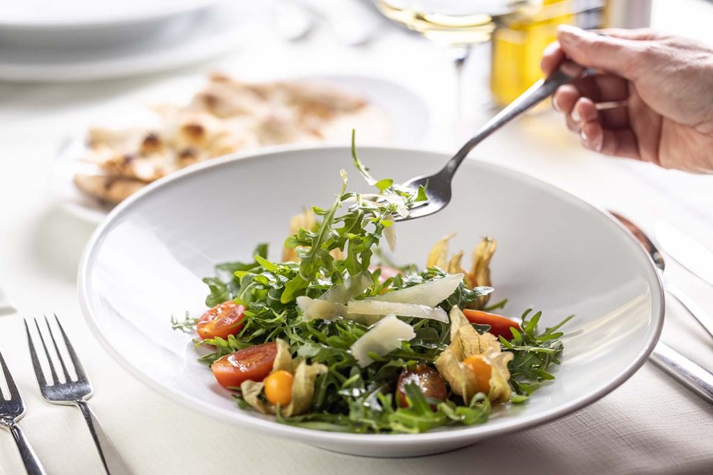 A hand using a fork to serve arugula salad with cherry tomatoes and cheese in a white bowl.