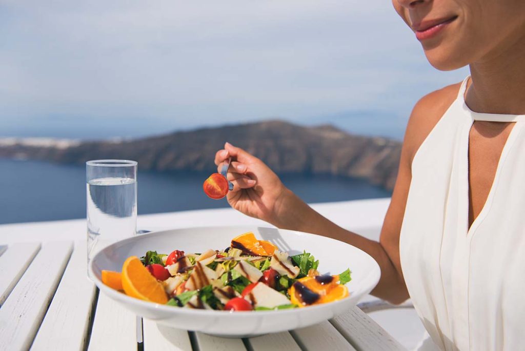 A woman holding a cherry tomato while enjoying a salad with oranges and greens, with a glass of water nearby.