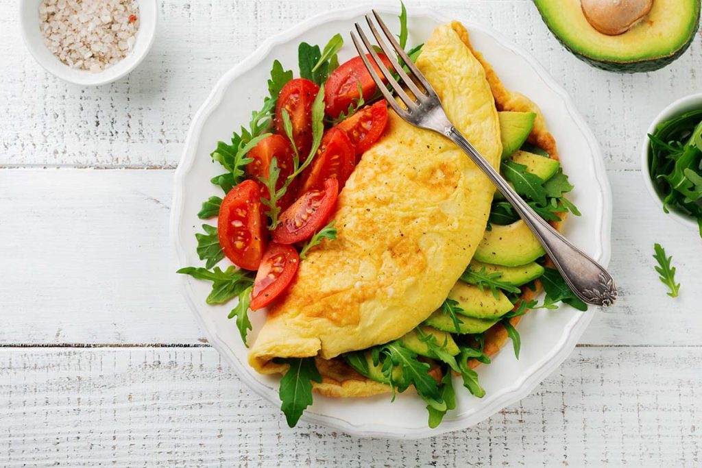 Omelette with avocado slices and cherry tomatoes served on a white plate with arugula. Fresh ingredients arranged neatly.