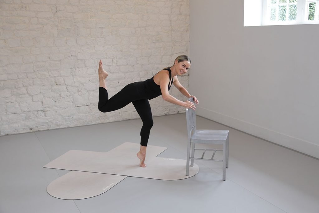A woman in black activewear balancing on one leg while holding onto a chair for support.