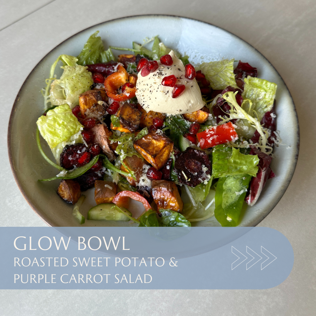 Salad bowl containing roasted sweet potato, purple carrot, mixed greens, and garnished with pomegranate seeds and a dollop of cream.