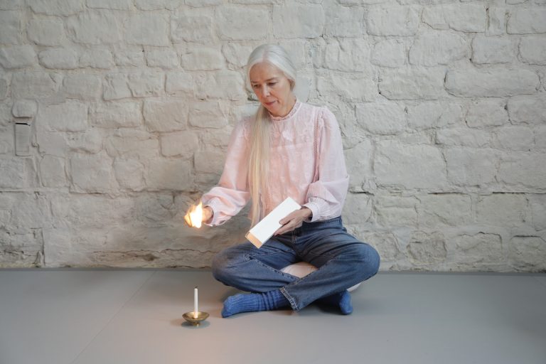 A woman with long grey hair sitting on the floor, holding a book and lighting a candle with a flame.