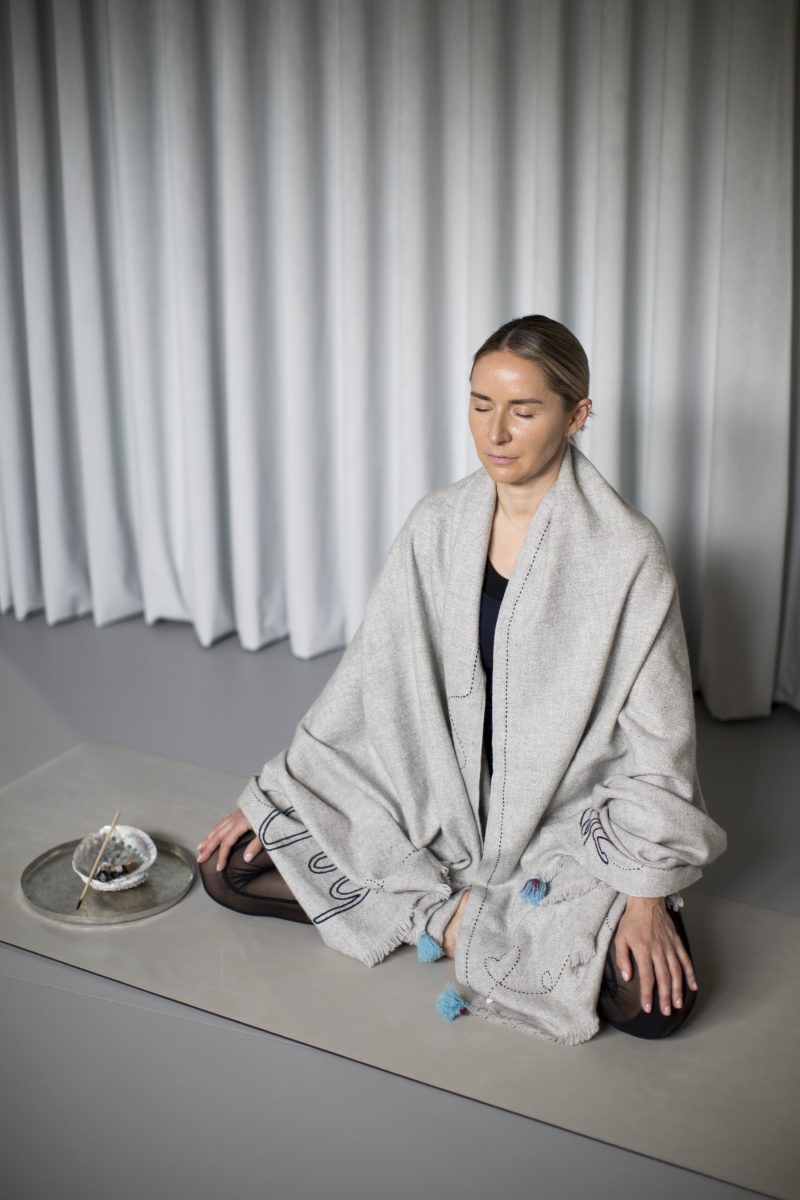 Woman meditating while wrapped in a grey shawl, seated on a smooth surface with a decorative plate nearby.