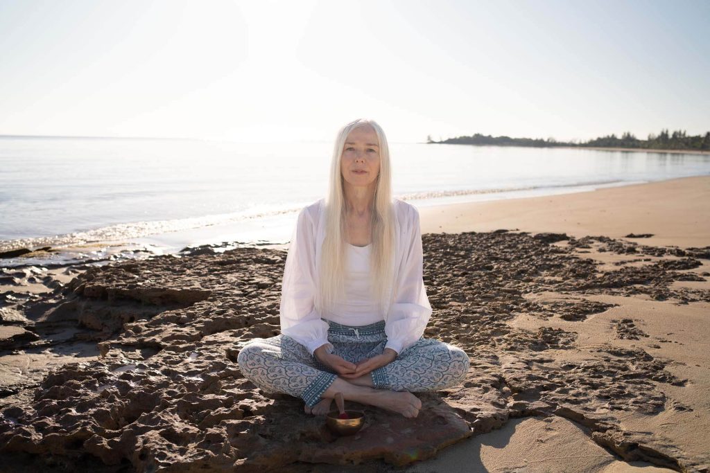 Woman with long white hair meditating on a rocky beach by the sea, wearing a white top and patterned trousers.