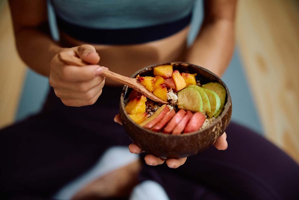 A bowl containing granola topped with slices of peach, watermelon, and lime held by a person sitting on a yoga mat.