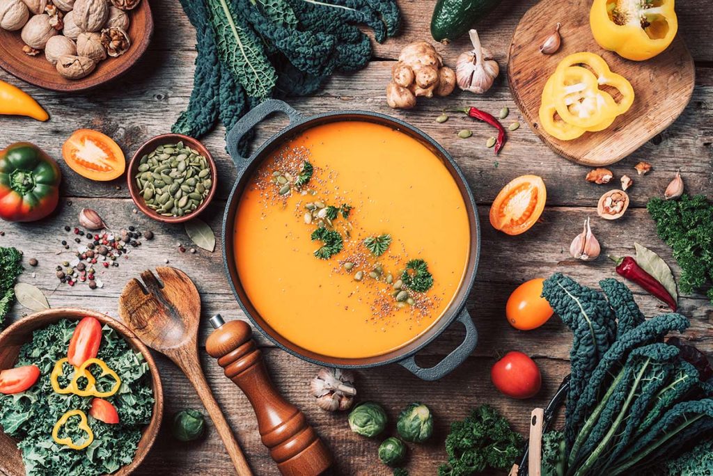 A bowl of orange vegetable soup garnished with herbs, surrounded by fresh vegetables and spices on a wooden surface.