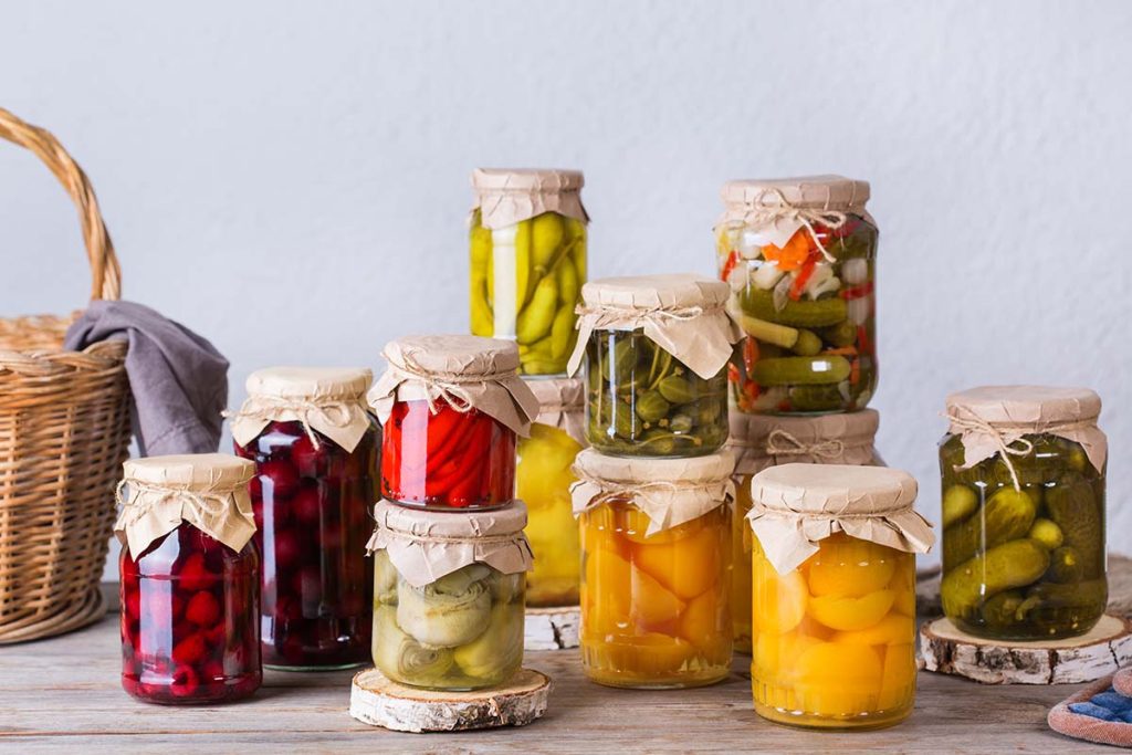 Various jars containing preserved vegetables and fruits, including pickles and fruits in syrup, arranged on a wooden surface.