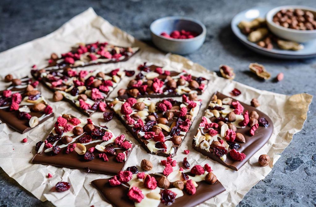 Chocolate bark topped with nuts, dried raspberries, and cranberries arranged on parchment paper.
