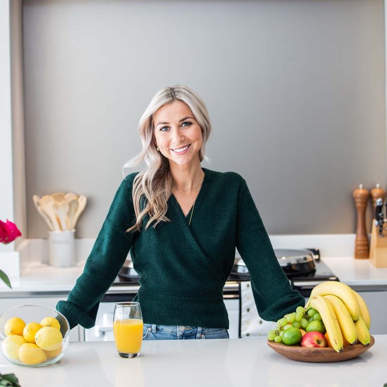 A woman in a green sweater standing at a kitchen counter with a bowl of fruit and a glass of orange juice.