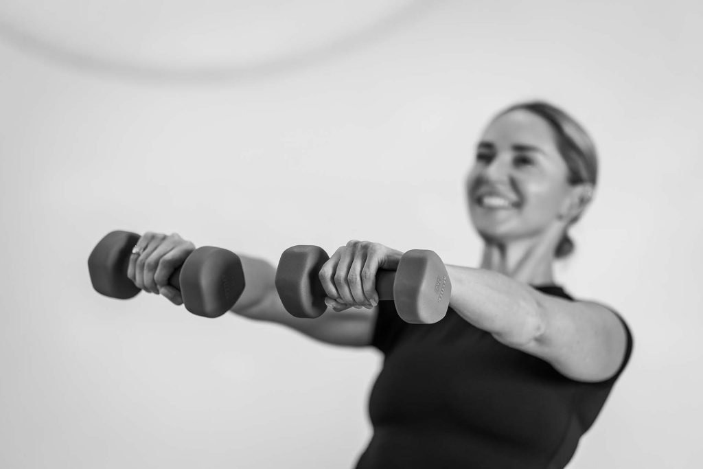 A woman smiling while holding two dumbbells in a workout position, demonstrating strength training.