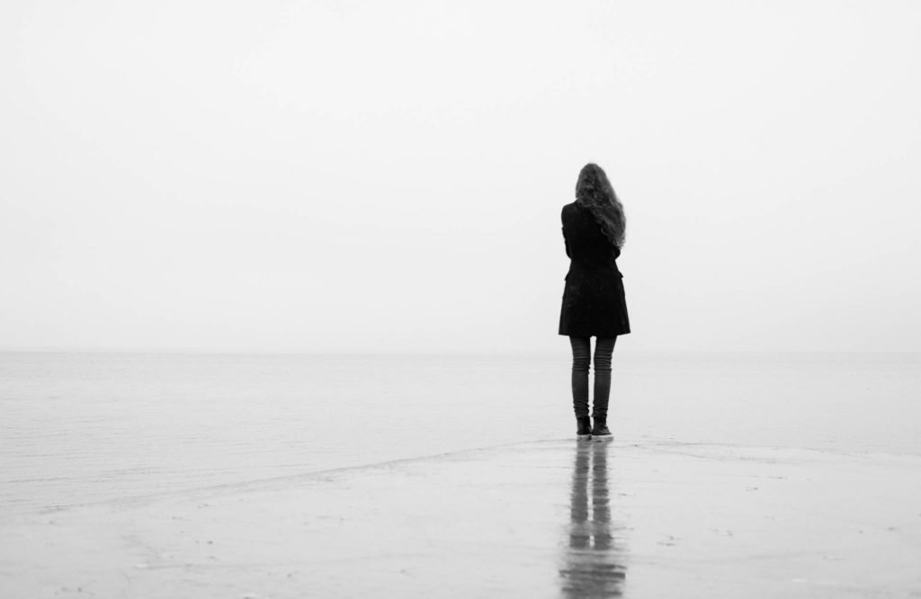 A woman with long hair wearing a black coat stands at the edge of a body of water, reflecting on the surface.