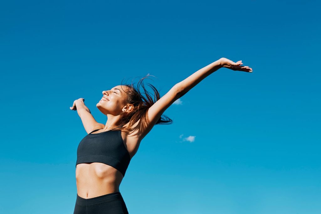 Woman in a black crop top with arms raised, smiling against a clear blue sky.
