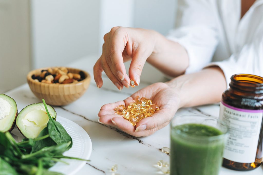 Person holding dietary supplements in one hand, with spinach, avocado, nuts, and a green smoothie on a table.