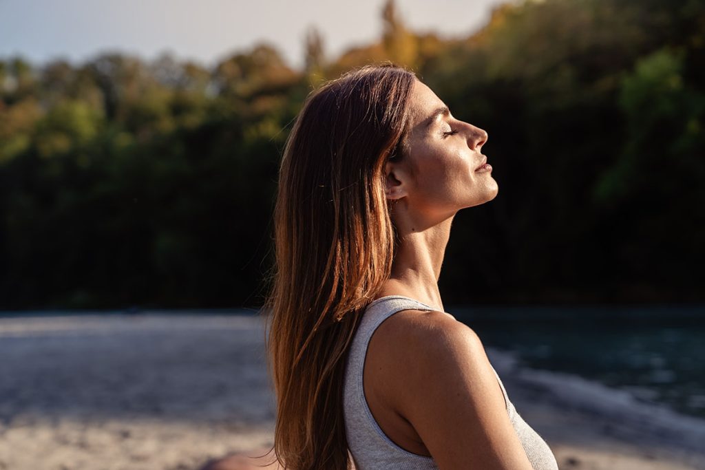 Woman with long brown hair standing in profile, eyes closed, enjoying a moment of calm outdoors.