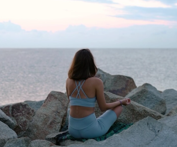 Individual in grey activewear meditating on rocks by the sea during sunset.