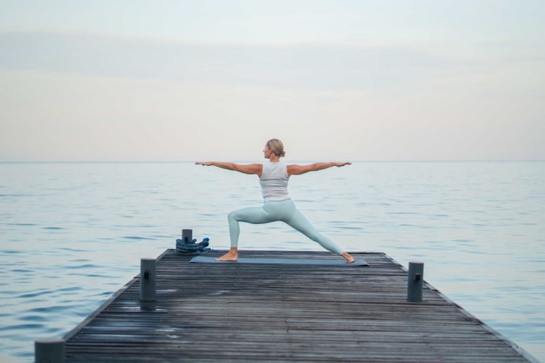A woman performing a yoga pose on a wooden pier extending over calm water.