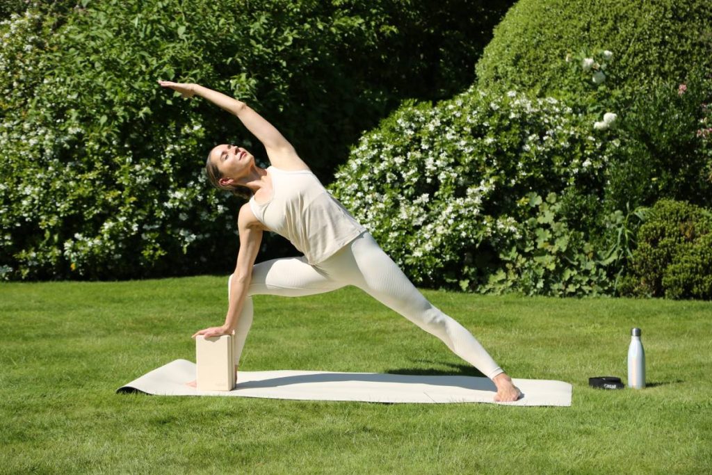 A woman performing a yoga pose with a block on a mat in a green outdoor setting.