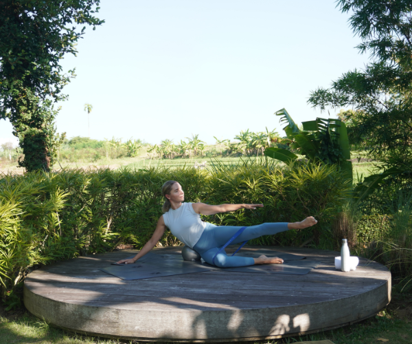 Woman in activewear performing a side leg lift exercise on a wooden platform surrounded by greenery.