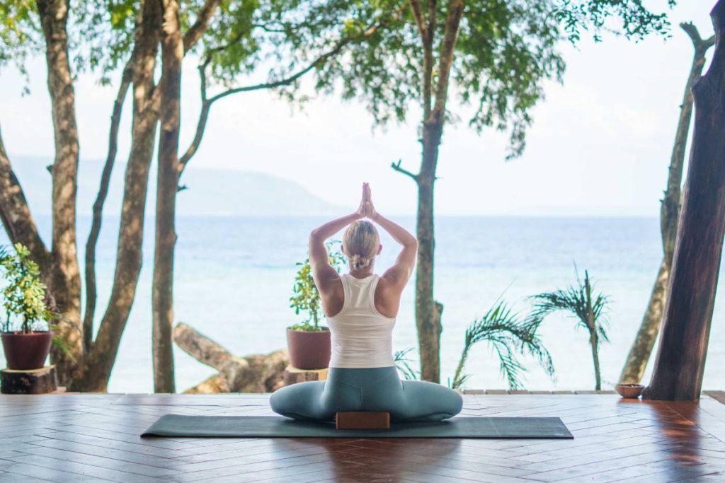 Individual performing a seated yoga pose on a mat with hands raised in prayer position, overlooking the sea.