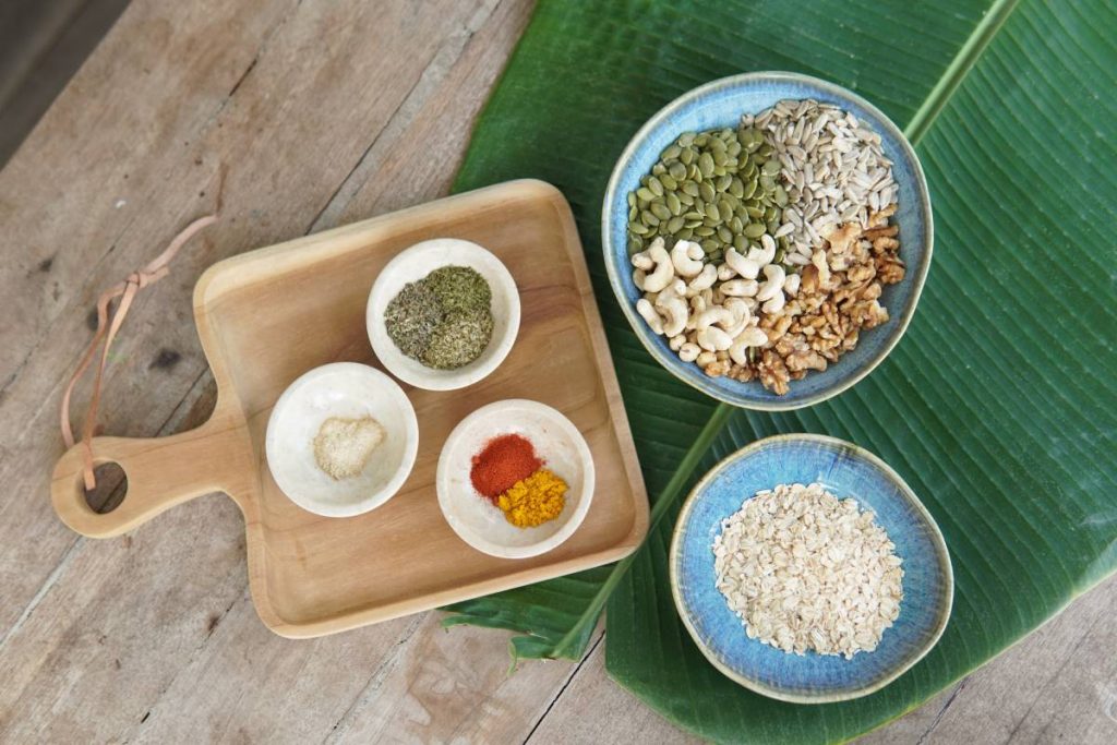 Various ingredients for savoury nutty granola displayed in bowls, including seeds, nuts, and spices on a wooden surface.