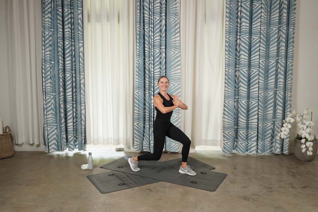 A woman performing a lunge exercise on a mat in a well-lit studio with patterned curtains.