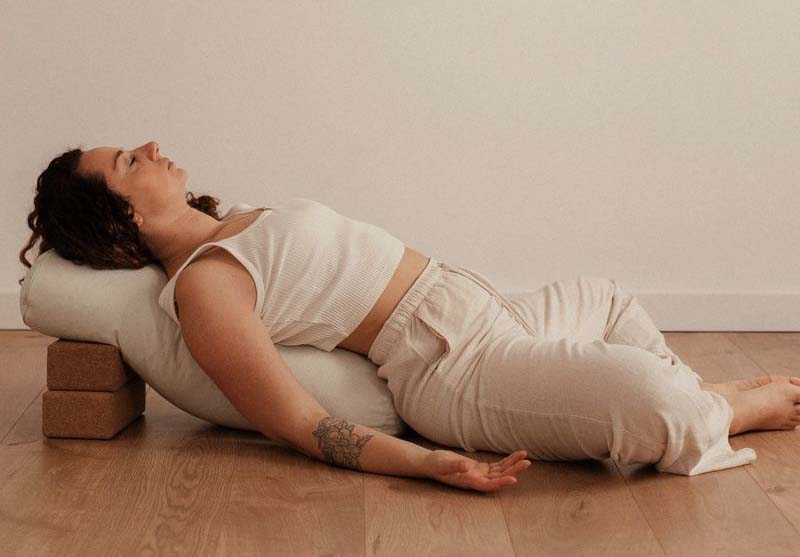 Individual in a restorative yoga pose using blocks and a bolster for support on a wooden floor.