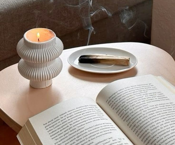 A white ribbed candle holder with a lit candle beside a plate containing a smudging stick and an open book.